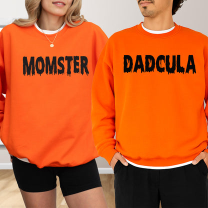 May include: Two orange sweatshirts with black text. One says "MOMSTER" and the other says "DADCULA". Both sweatshirts have a white crew neck collar. The sweatshirts are worn with black shorts and pants.