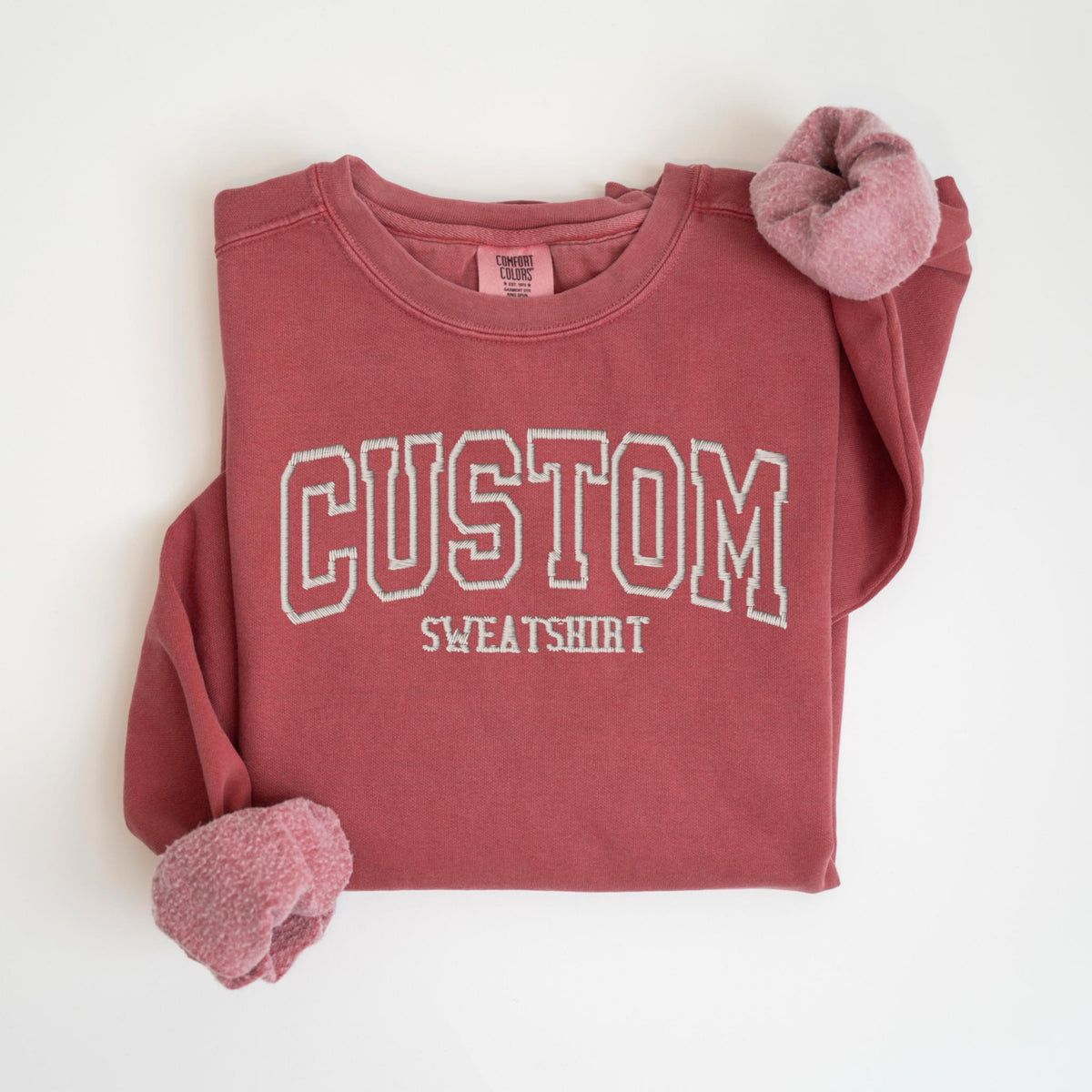 May include: A maroon-colored custom sweatshirt with the word "CUSTOM" embroidered in white block letters. The word "SWEATSHIRT" is embroidered below. The sleeves have a soft, pinkish-red lining. The sweatshirt is laid flat on a white surface.