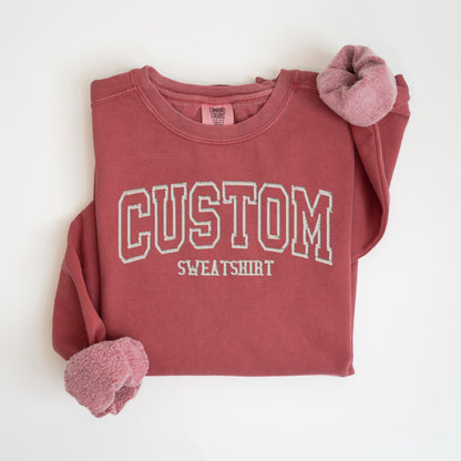 May include: A maroon-colored custom sweatshirt with the word "CUSTOM" embroidered in white block letters. The word "SWEATSHIRT" is embroidered below. The sleeves have a soft, pinkish-red lining. The sweatshirt is laid flat on a white surface.