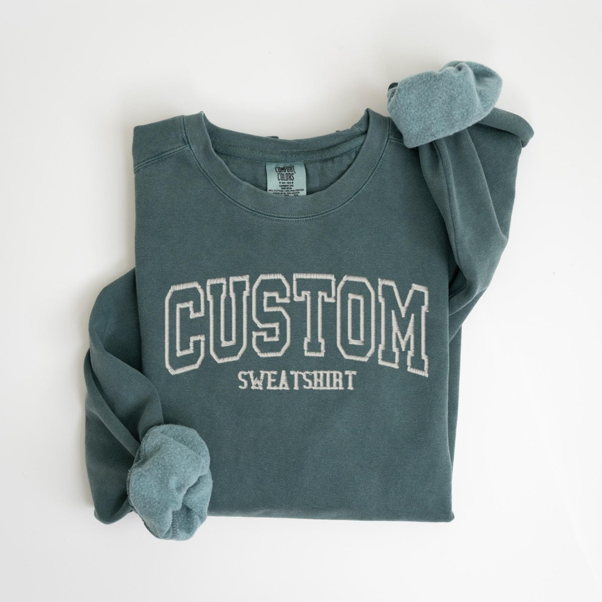 May include: A teal-colored sweatshirt with the word "CUSTOM" embroidered in white block letters. The word "SWEATSHIRT" is embroidered below. The sleeves are cuffed, and the garment is folded on a white surface.