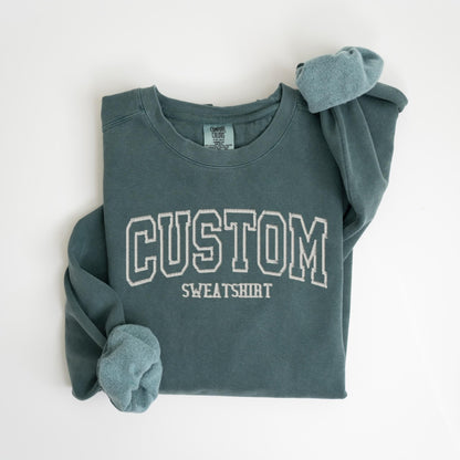 May include: A teal-colored sweatshirt with the word "CUSTOM" embroidered in white block letters. The word "SWEATSHIRT" is embroidered below. The sleeves are cuffed, and the garment is folded on a white surface.