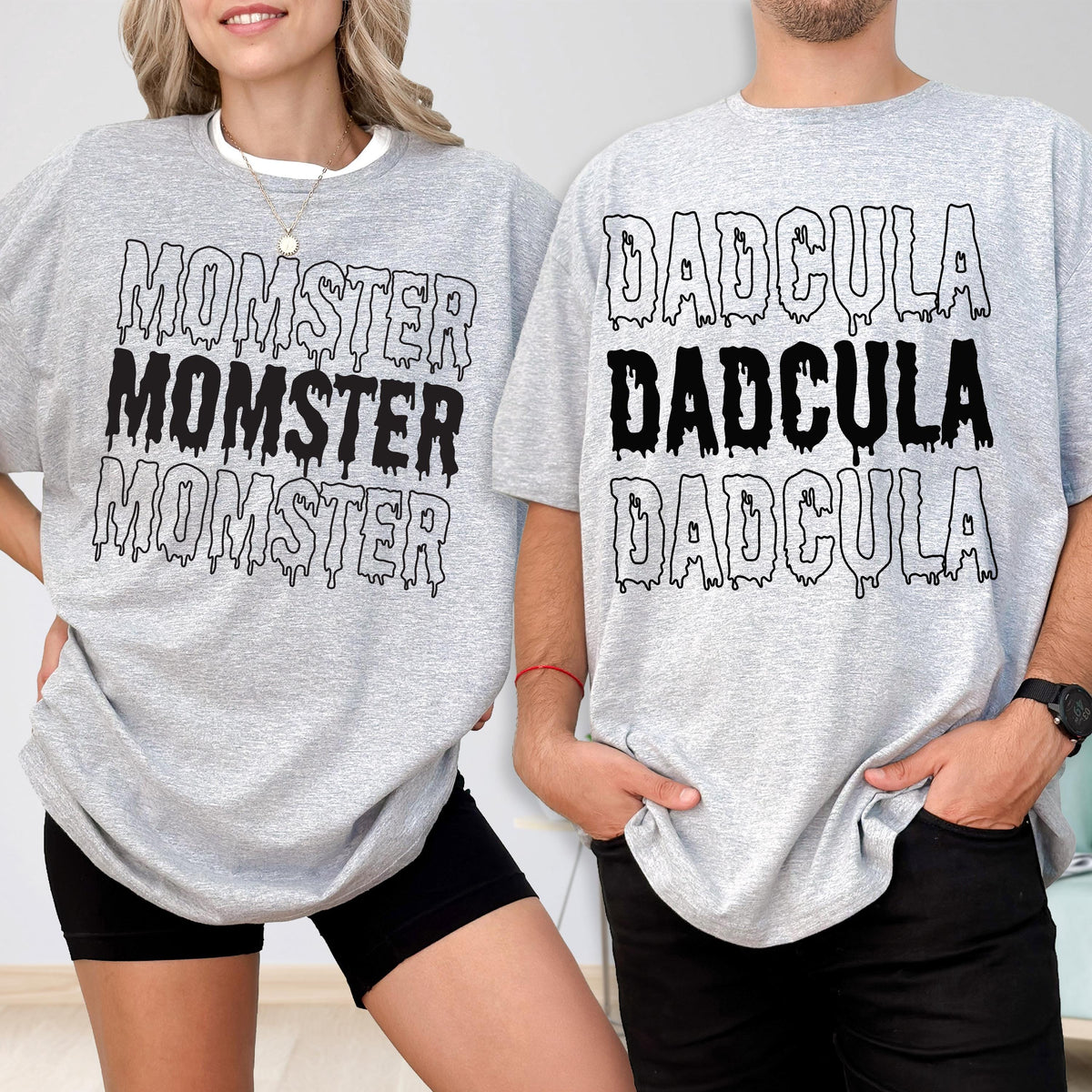 May include: Two gray t-shirts with black text. One shirt says "MOMSTER" three times, the other says "DADcula" three times. The shirts are worn by two people. The shirts are a Halloween theme.