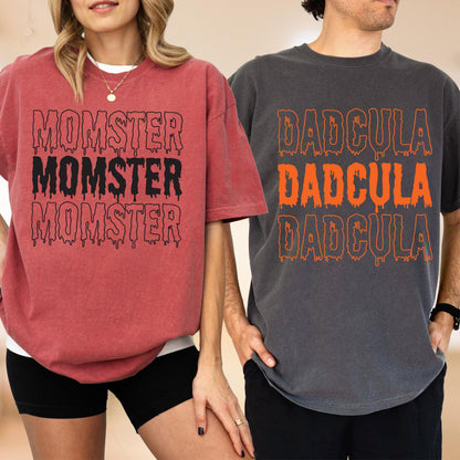 May include: Two short-sleeved t-shirts. One is a reddish-pink color with the word "MOMSTER" in black dripping font. The other is a dark gray color with the word "DADcula" in orange dripping font. Both shirts are oversized.