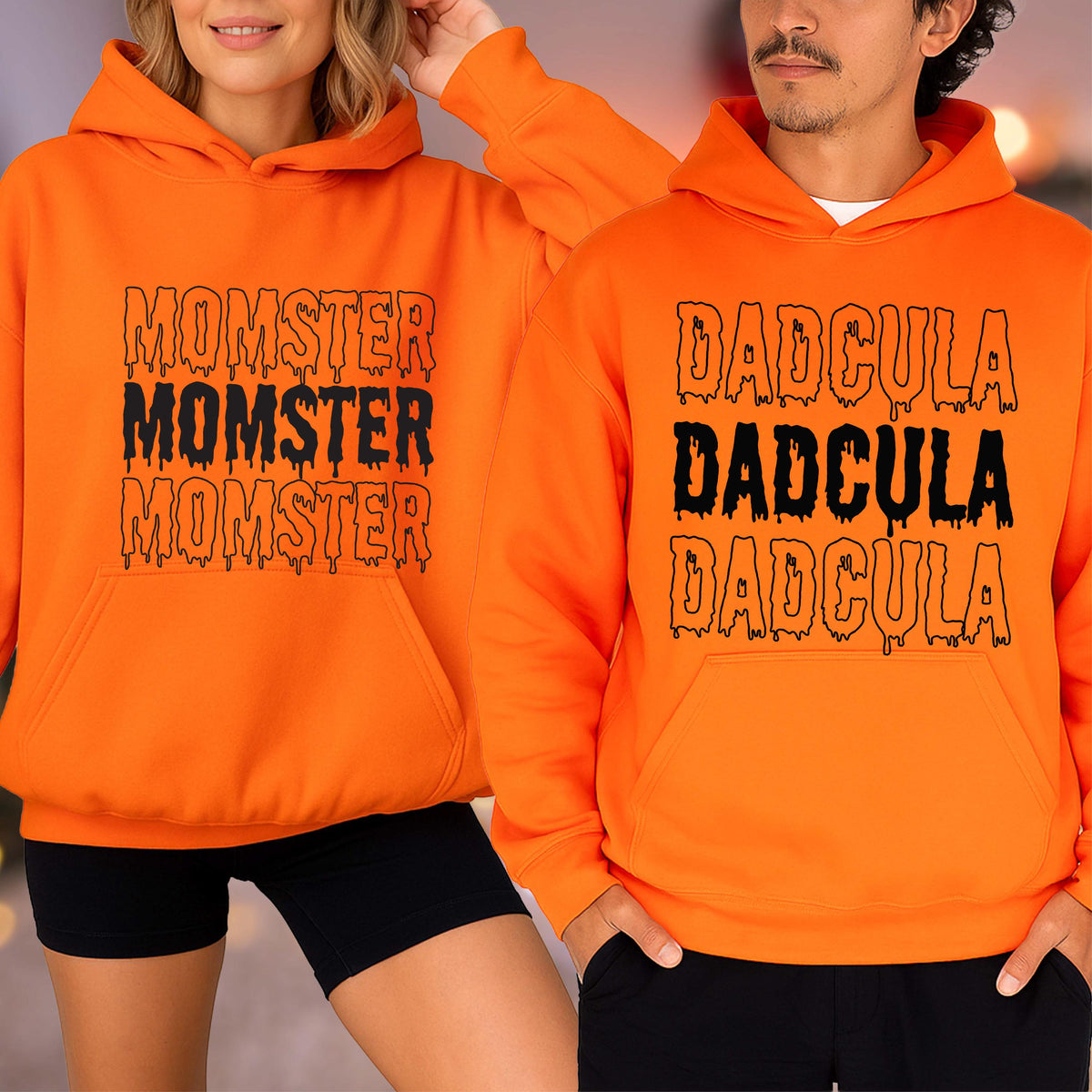 May include: Two orange hoodies with black text. One hoodie says "MOMSTER" three times, and the other says "DADcula" three times. Both hoodies have a front pocket and a hood. The hoodies are worn by two people.