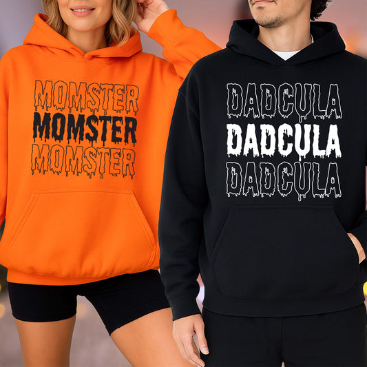 May include: Two hooded sweatshirts, one orange and one black. The orange sweatshirt has the text "MOMSTER MOMSTER MOMSTER" in black dripping font. The black sweatshirt has the text "DADGULA DADGULA DADGULA" in white dripping font.