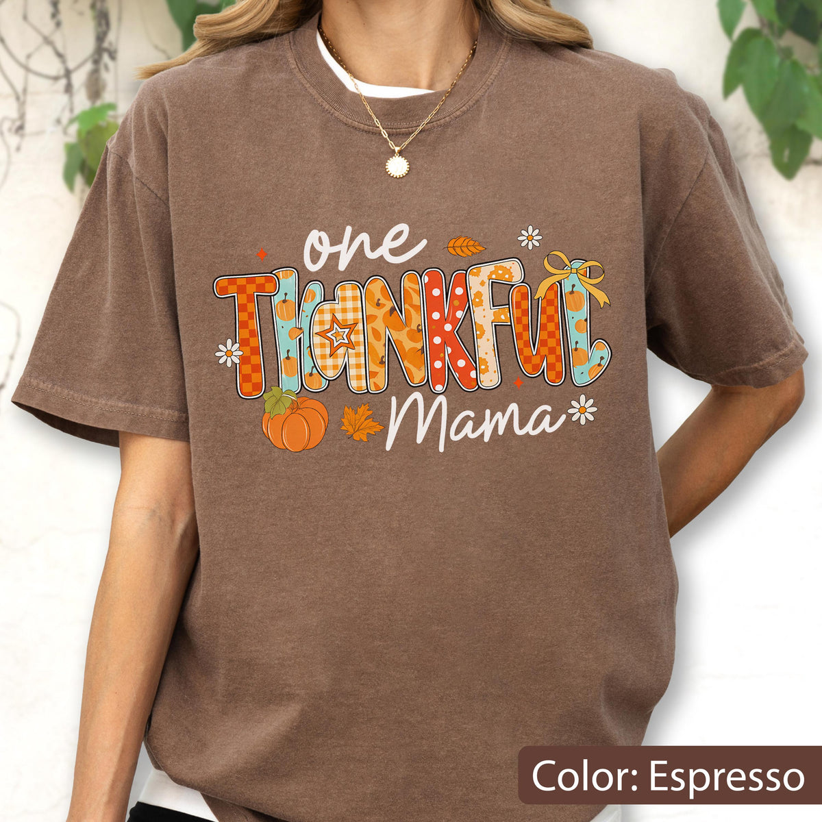 May include: Espresso-colored t-shirt with the words "one THANKFUL Mama" in a colorful, fall-themed design. The design includes pumpkins, leaves, and floral accents. The shirt has short sleeves and a crew neck.