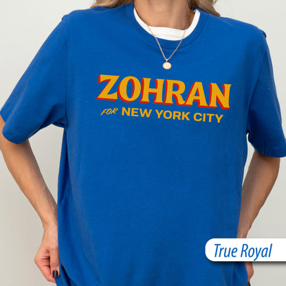 May include: A true royal blue short-sleeved t-shirt featuring the yellow text ZOHRAN FOR NEW YORK CITY with a red drop shadow effect. The shirt has a crew neckline and is worn over a white undershirt, paired with a thin gold chain necklace.
