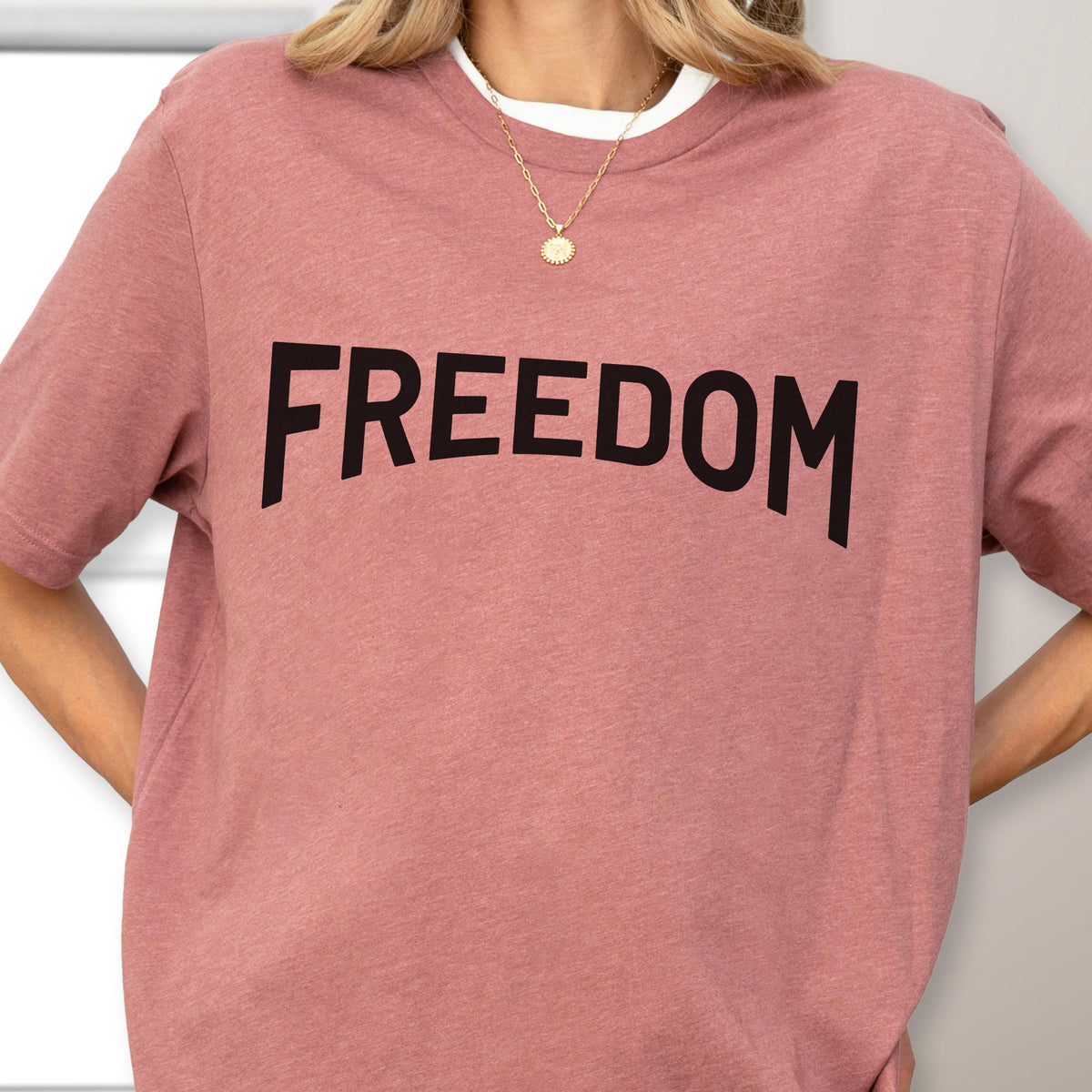 May include: A dusty rose short-sleeve t-shirt with the word "FREEDOM" printed in bold black letters across the front. The shirt has a white crew neck collar peeking out from the neckline. The shirt is made of a soft, comfortable material.