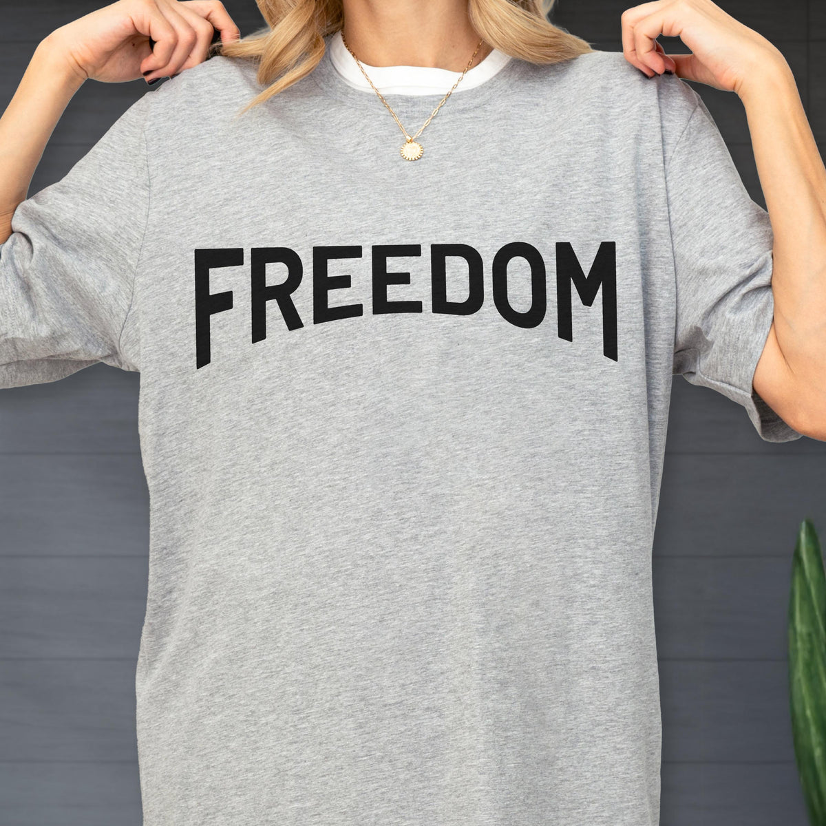 May include: A heather gray t-shirt with the word "FREEDOM" printed in bold, black letters across the front. The shirt has short sleeves and a crew neck, with a white collar peeking out. A gold necklace is visible.