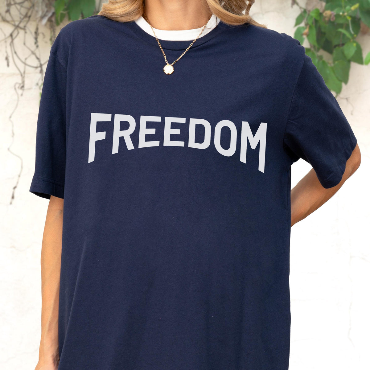 May include: Navy blue t-shirt with the word "FREEDOM" in large white capital letters. The shirt has a white collar and is worn with a gold necklace.