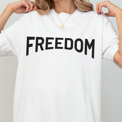 May include: White t-shirt with the word "FREEDOM" printed in large, black, bold letters across the front. The shirt has short sleeves and a crew neck. The overall style is casual and simple.