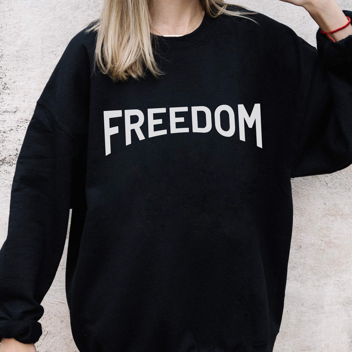 May include: Black sweatshirt with the word "FREEDOM" printed in large, white, block letters across the chest. The sweatshirt has a crew neck and long sleeves. The background is a light gray wall.
