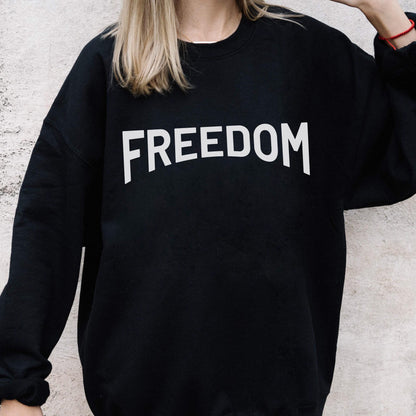 May include: Black sweatshirt with the word "FREEDOM" printed in large, white, block letters across the chest. The sweatshirt has a crew neck and long sleeves. The background is a light gray wall.