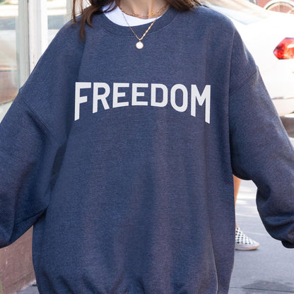 May include: A dark blue sweatshirt with the word "FREEDOM" printed in large, white capital letters across the chest. The sweatshirt has a crew neck and long sleeves. The person is wearing a white t-shirt underneath and a gold necklace.