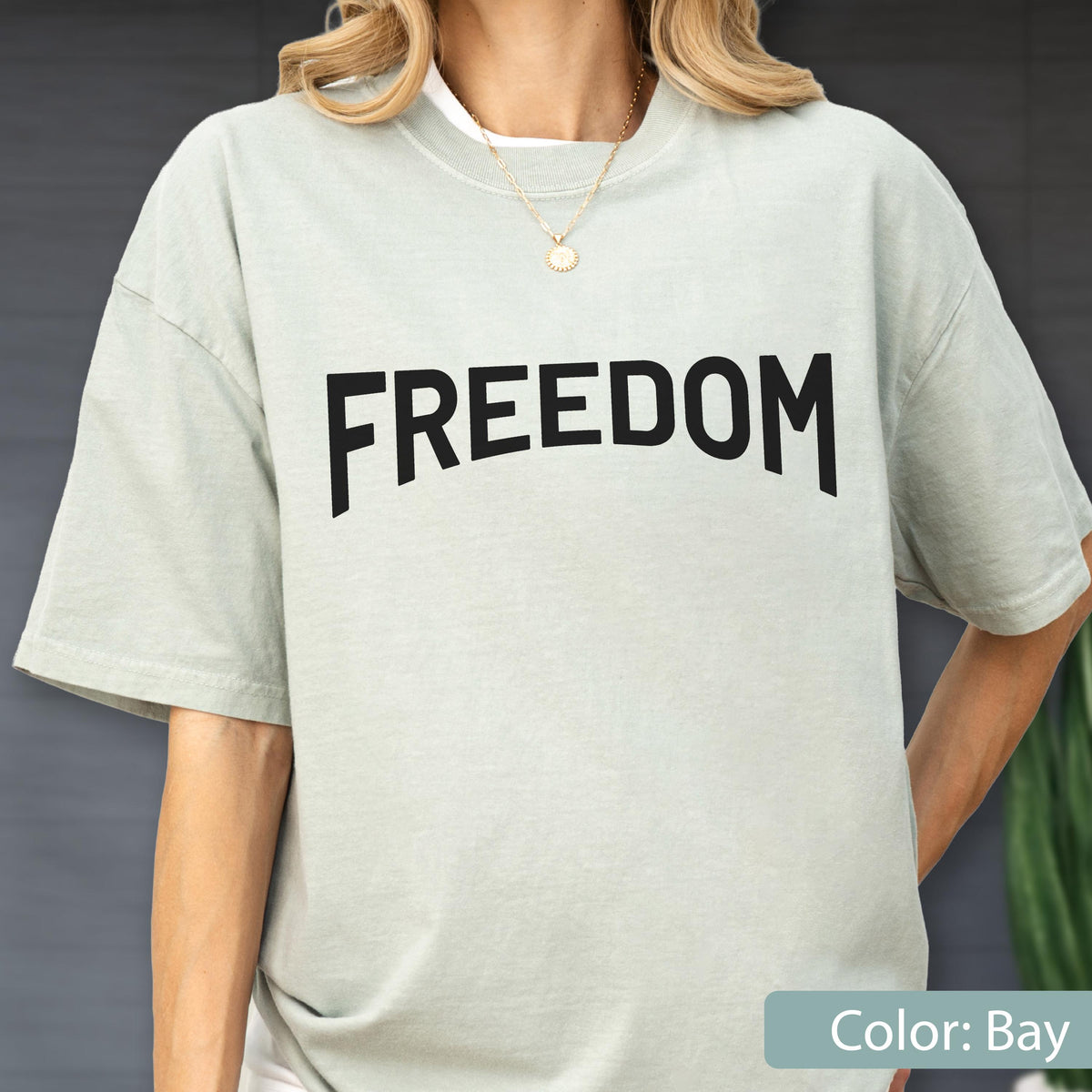 May include: A light green t-shirt with the word "FREEDOM" printed in bold black letters. The shirt has short sleeves and a crew neck. The color of the shirt is "Bay".