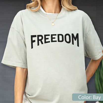 May include: A light green t-shirt with the word "FREEDOM" printed in bold black letters. The shirt has short sleeves and a crew neck. The color of the shirt is "Bay".
