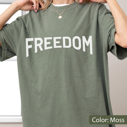 May include: A moss green t-shirt with the word "FREEDOM" printed in large, white capital letters across the chest. The shirt has short sleeves and a crew neck. The color is described as "Moss".