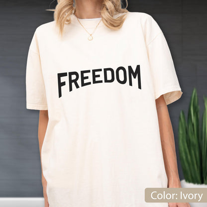 May include: Ivory-colored t-shirt with the word "FREEDOM" printed in bold, black letters across the front. The shirt has short sleeves and a relaxed fit. The color is ivory.