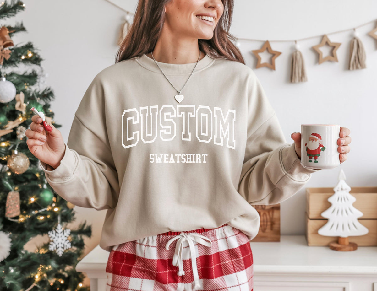 May include: A beige sweatshirt with the word "CUSTOM SWEATSHIRT" in white block letters. The person is wearing red and white plaid pajama pants and holding a mug with a Santa Claus graphic.