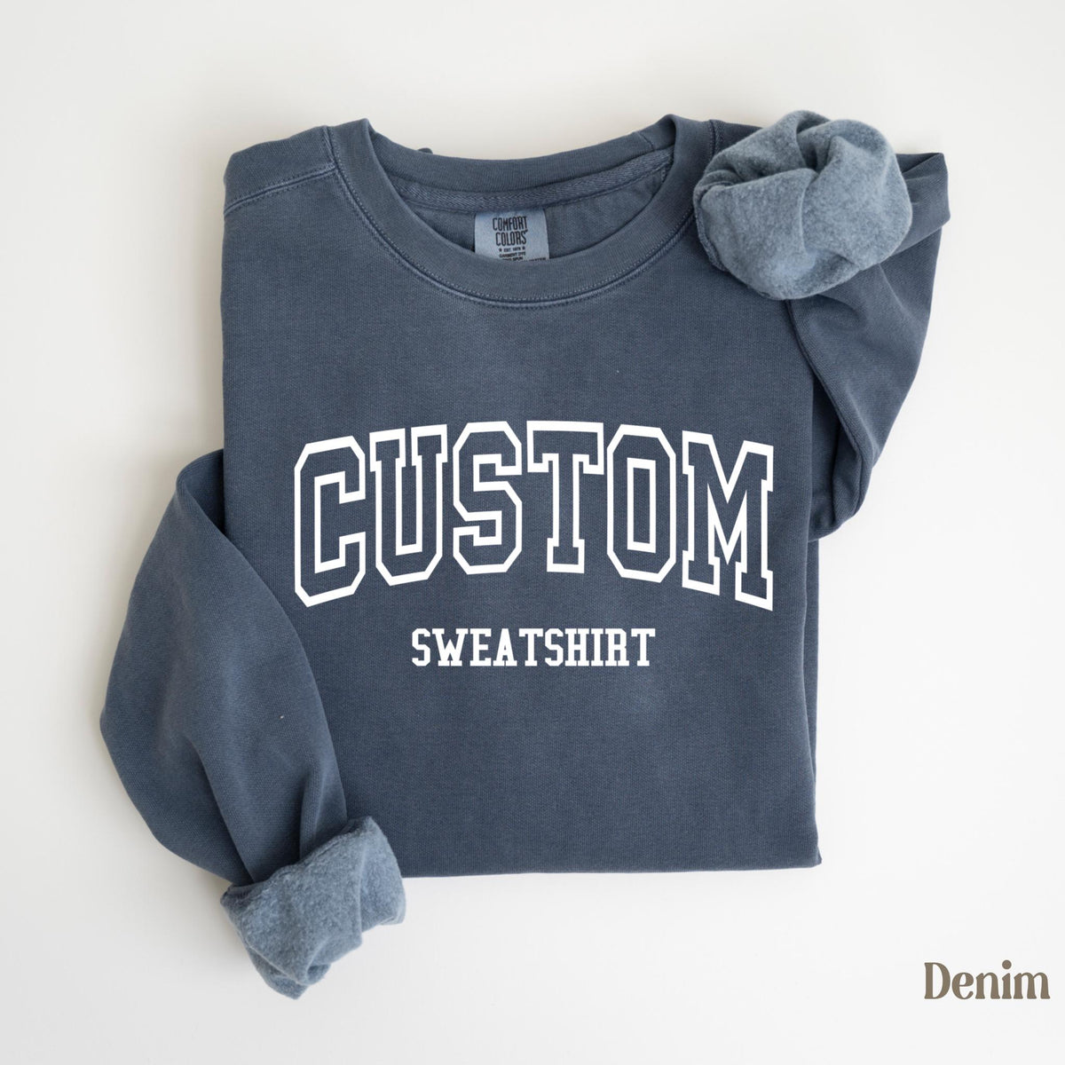 May include: A denim blue sweatshirt with the word "CUSTOM SWEATSHIRT" printed in white block letters. The sleeves are folded, revealing a lighter blue interior. The sweatshirt is laid flat on a white surface.