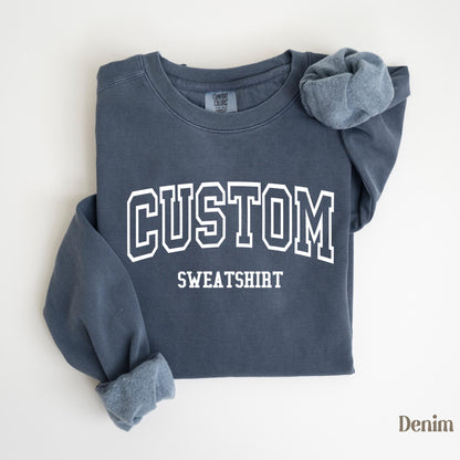 May include: A denim blue sweatshirt with the word "CUSTOM SWEATSHIRT" printed in white block letters. The sleeves are folded, revealing a lighter blue interior. The sweatshirt is laid flat on a white surface.