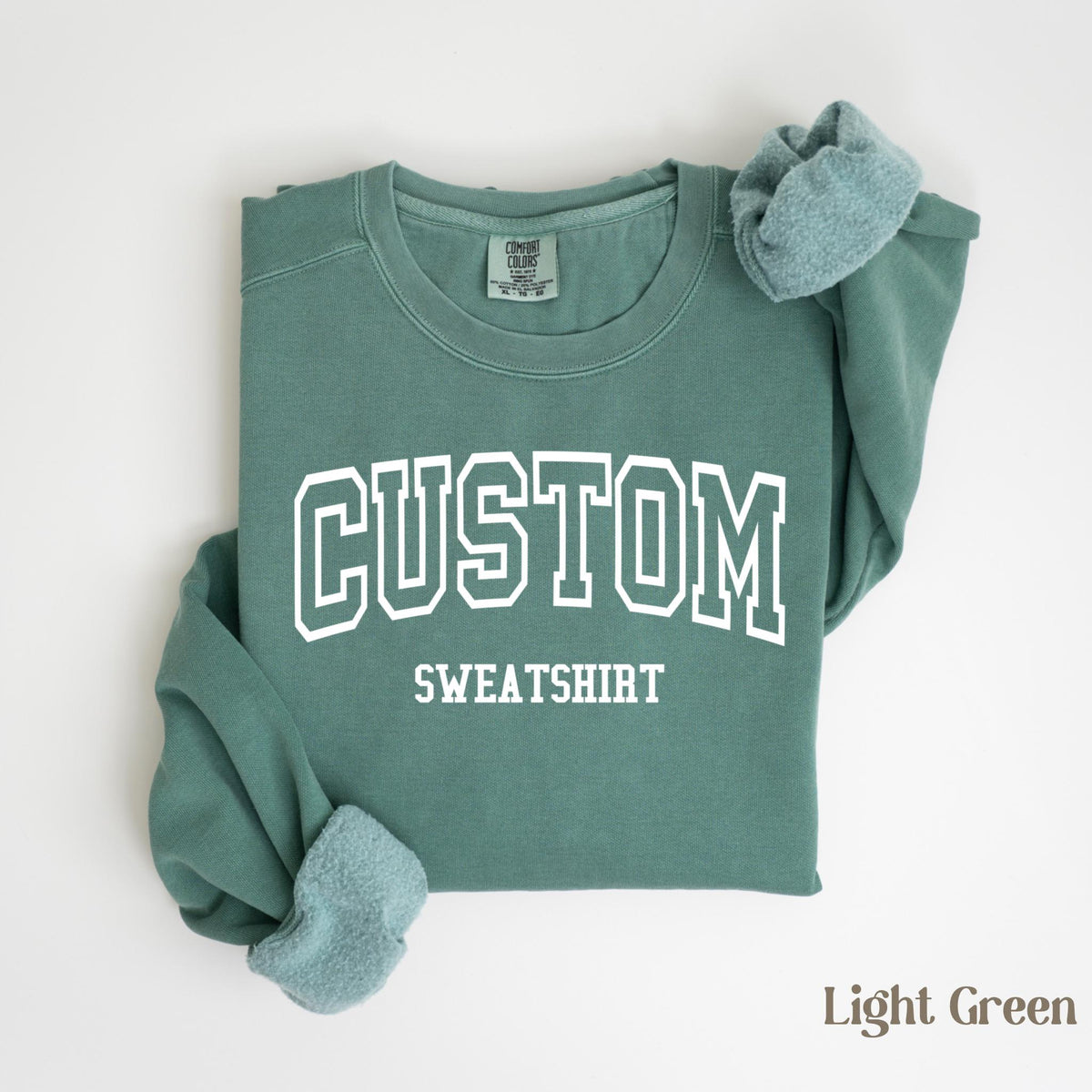 May include: Light green sweatshirt with the word "CUSTOM" in white block letters above the word "SWEATSHIRT". The sweatshirt has a crew neck and long sleeves, with the cuffs folded over. The words "Comfort Colors" are printed on the tag.