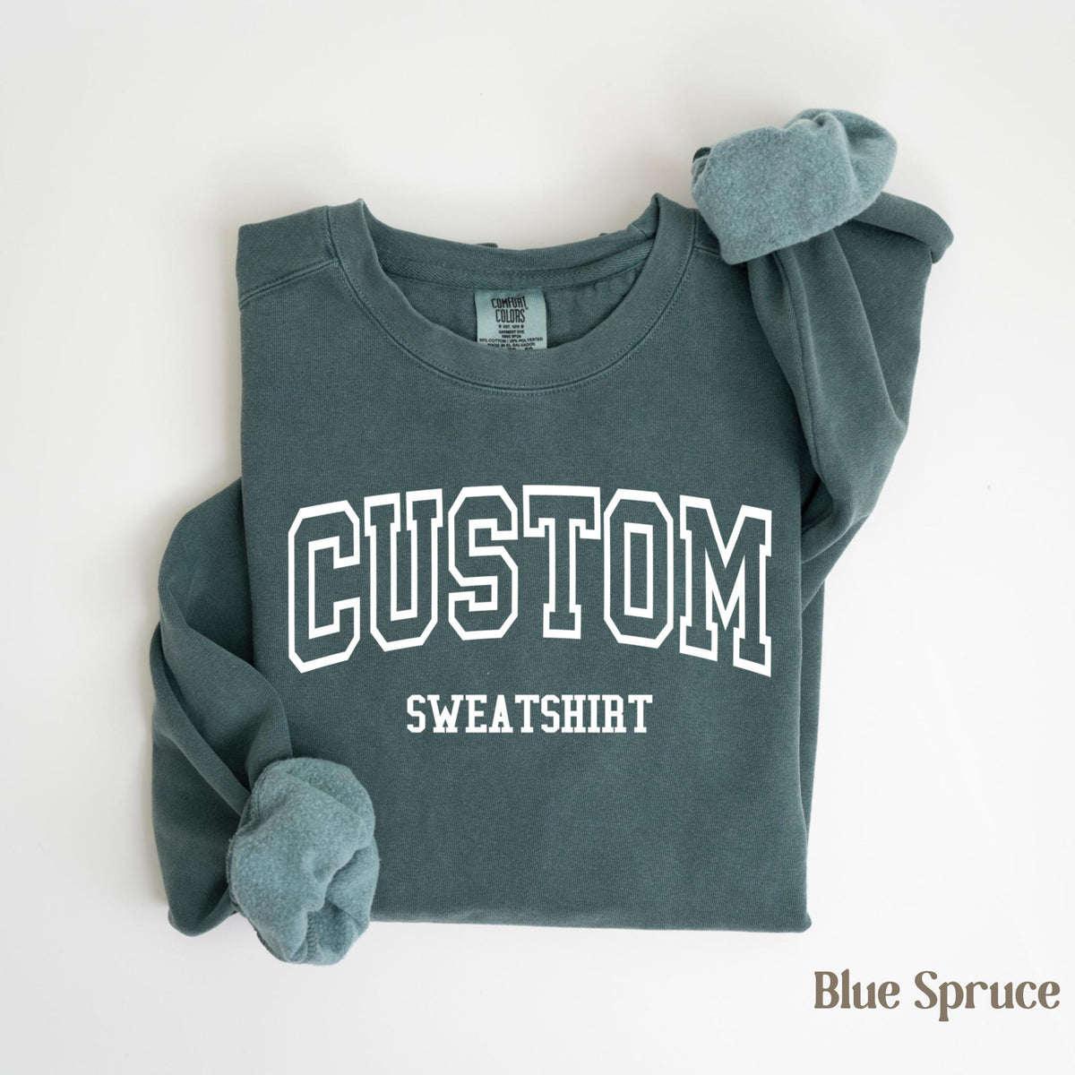 May include: A blue spruce sweatshirt with the word "CUSTOM SWEATSHIRT" printed in white block letters. The sleeves are cuffed, and the sweatshirt is folded neatly. The words "Blue Spruce" are printed in the bottom right corner.