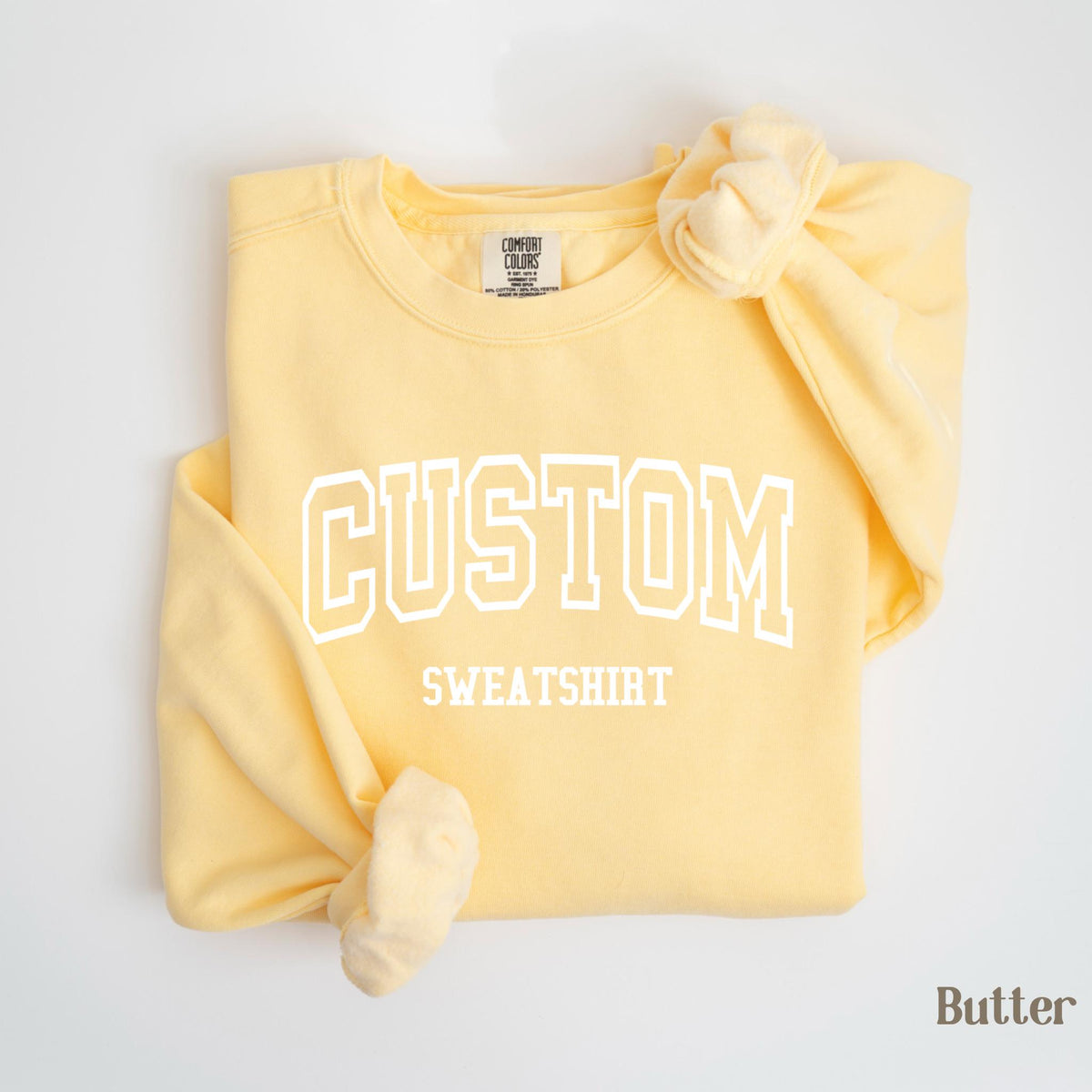 May include: A light yellow sweatshirt with the word "CUSTOM" in white block letters above the word "SWEATSHIRT". The sleeves are knotted. The sweatshirt is labeled "Butter".
