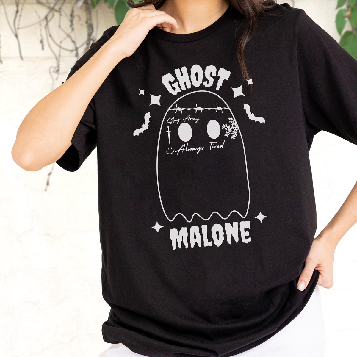 May include: A black t-shirt featuring a white graphic of a ghost with the text GHOST MALONE. The ghost design includes a barbed wire crown, a sword, a snowflake, a smiley face, and the text Stay Away and Always Tired, surrounded by stars and bats.