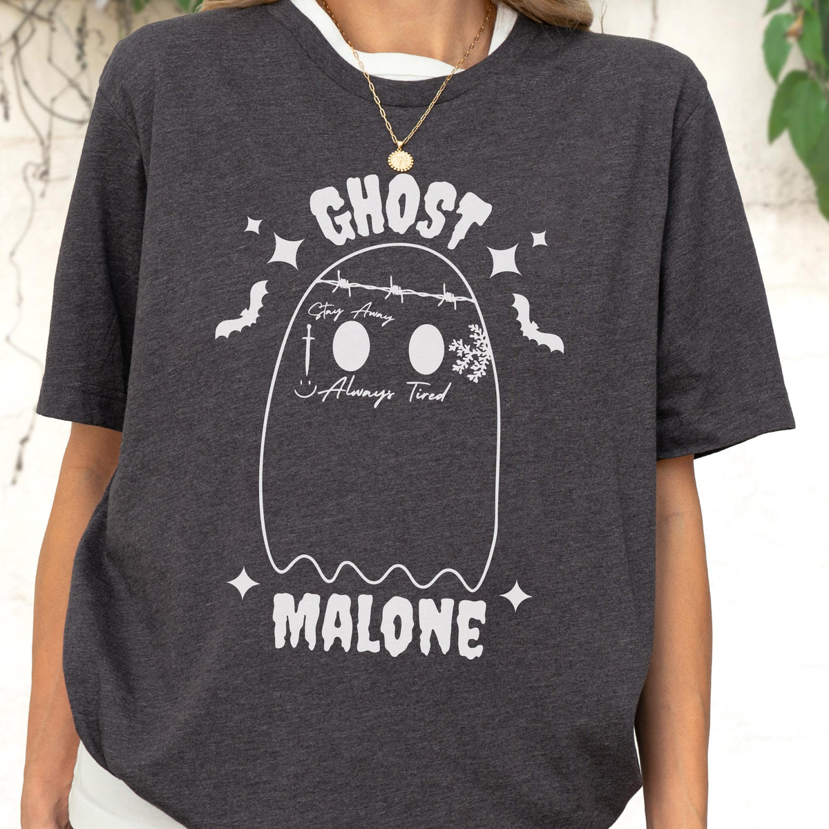 May include: A charcoal gray short-sleeve t-shirt featuring a white graphic of a ghost with barbed wire, a sword, a snowflake, and the text "GHOST MALONE," "Stay Away," and "Always Tired." The design includes small stars and bat silhouettes.