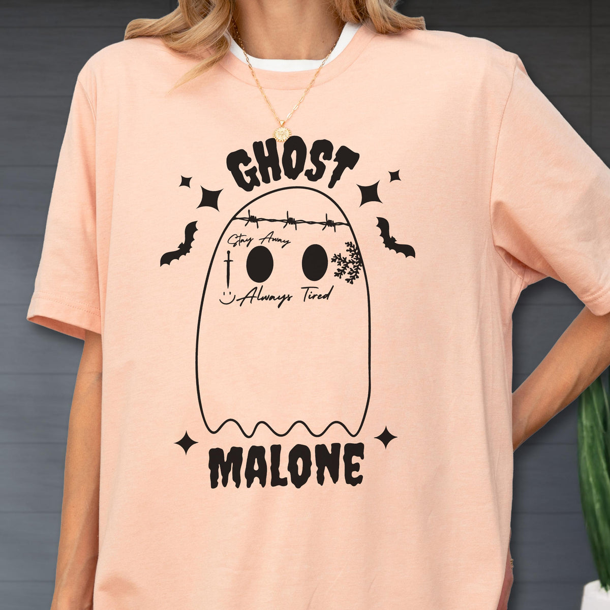 May include: A peach-colored short-sleeve t-shirt featuring a black graphic of a ghost with barbed wire across its head, a sword, a snowflake, and the text "GHOST MALONE", "Stay Away", and "Always Tired" printed in a bold, stylized font.