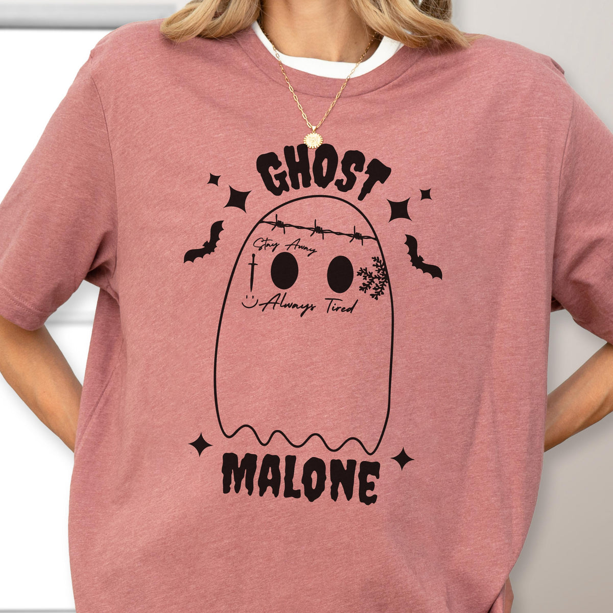 May include: A dusty rose t-shirt featuring a black graphic of a ghost with barbed wire across its head. The ghost has the text Stay Away, a sword, a smiley face, and Always Tired printed on it. The word GHOST is above and MALONE is below.