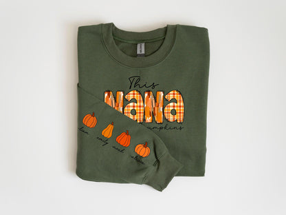 May include: Olive green sweatshirt with the text "This Nana" in black script and the word "Nana" in a plaid pattern. The sleeve features pumpkin illustrations and names. A fall-themed apparel item.