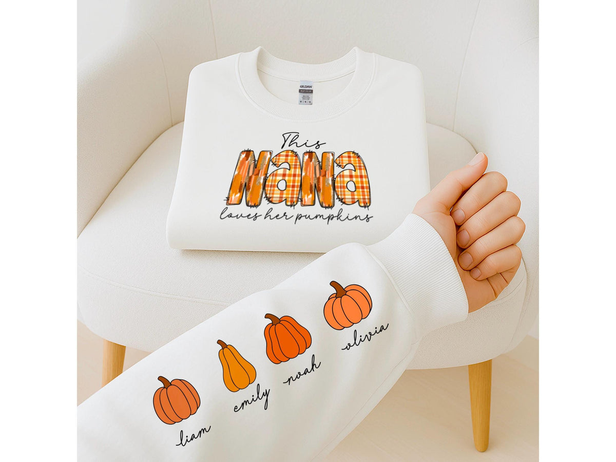 May include: A white sweatshirt with the text "This Nana loves her pumpkins" in orange plaid lettering. The sleeve features orange pumpkin illustrations with names: Liam, Emily, Noah, and Olivia. The sweatshirt is displayed on a white chair.