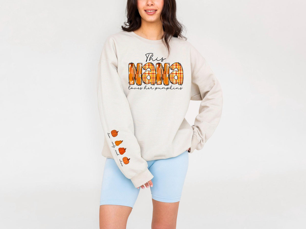 May include: Beige sweatshirt with the text "This Nana loves her pumpkins" in black script and orange plaid letters. The sleeve features pumpkin illustrations. The sweatshirt is paired with light blue shorts.