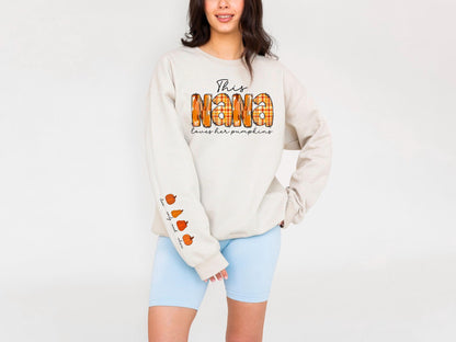 May include: Beige sweatshirt with the text "This Nana loves her pumpkins" in black script and orange plaid letters. The sleeve features pumpkin illustrations. The sweatshirt is paired with light blue shorts.