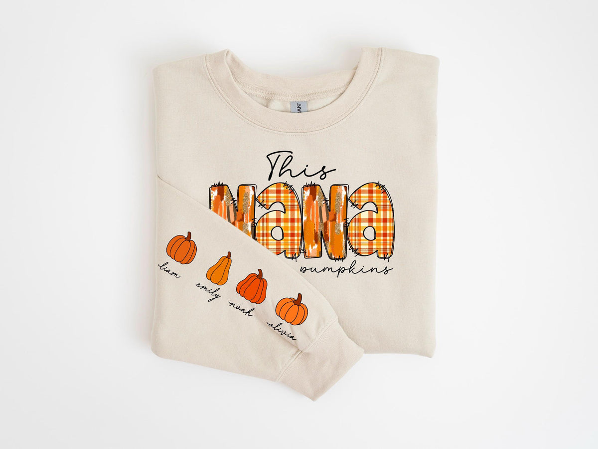 May include: Cream-colored sweatshirt with the text "This Nana loves pumpkins" in a mix of fonts and colors. The word "Nana" is in a plaid pattern. The left sleeve features pumpkin illustrations and names: Liam, Emily, Noah, and Olivia.