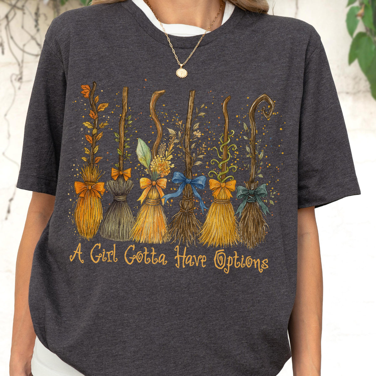 May include: A dark heather gray short-sleeve t-shirt featuring a graphic print of six distinct broomsticks adorned with colorful bows and foliage. Below the illustration, the text reads, A Girl Gotta Have Options in a stylized yellow cursive font.