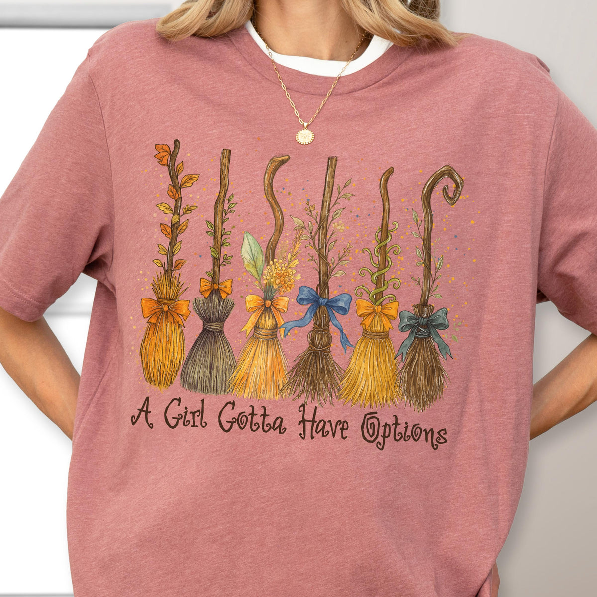 May include: A dusty rose short-sleeve t-shirt featuring a graphic of six distinct stylized brooms decorated with leaves, flowers, and ribbons. Below the brooms, the text reads, A Girl Gotta Have Options, in a dark brown cursive script font.