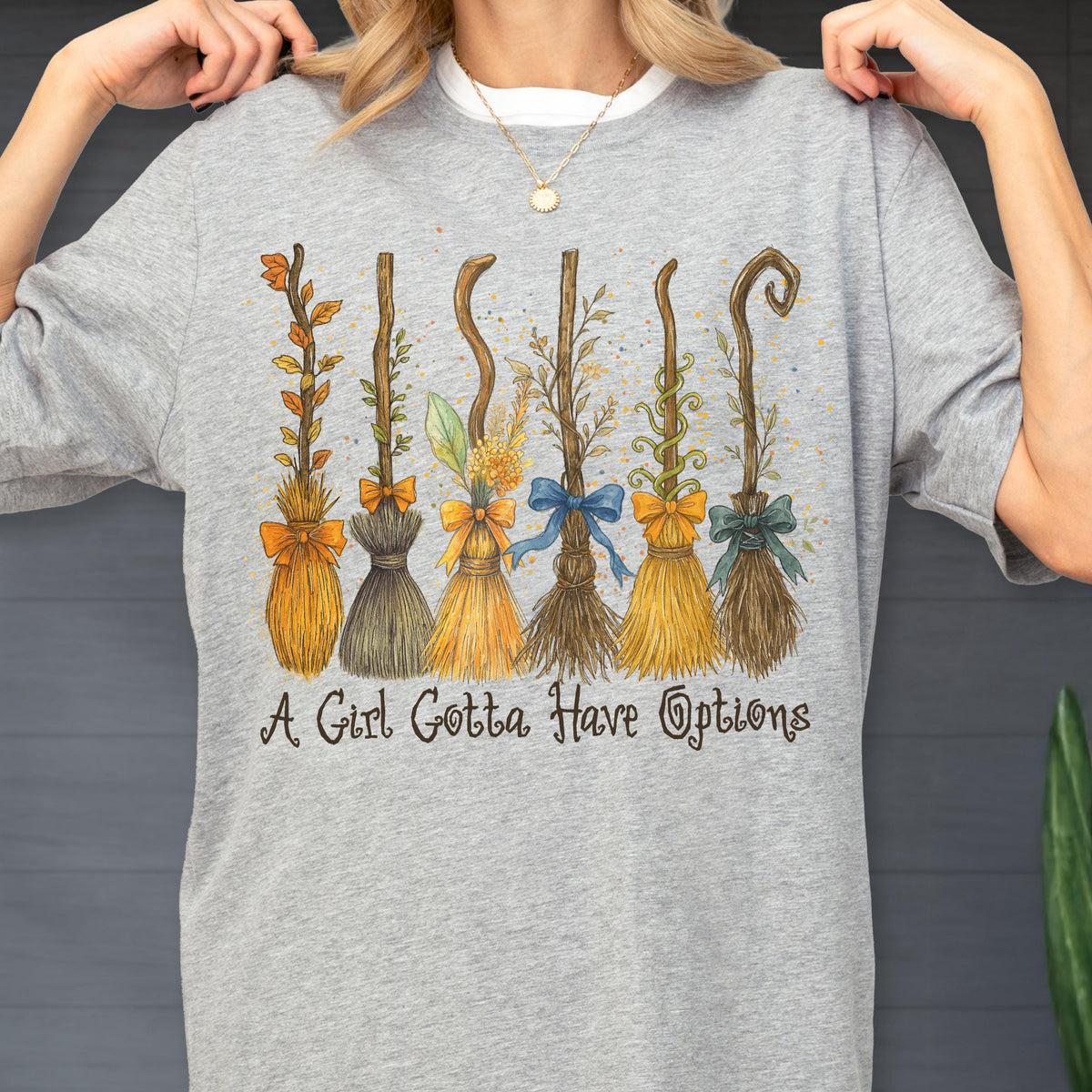 May include: A heather gray t-shirt featuring a graphic illustration of six distinct broomsticks decorated with various bows and foliage. Below the brooms, the text reads A Girl Gotta Have Options in a dark, stylized, cursive-inspired font.