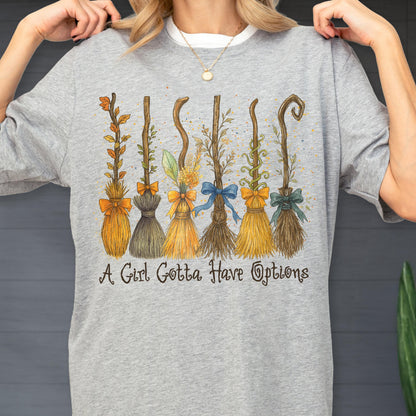 May include: A heather gray t-shirt featuring a graphic illustration of six distinct broomsticks decorated with various bows and foliage. Below the brooms, the text reads A Girl Gotta Have Options in a dark, stylized, cursive-inspired font.