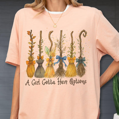 May include: A peach-colored t-shirt featuring a graphic of six distinct decorative brooms adorned with various ribbons and foliage. Below the illustration, the text reads, A Girl Gotta Have Options, in a dark brown, stylized cursive font.