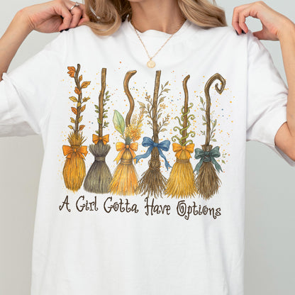 May include: A white short-sleeved t-shirt featuring a graphic of six distinct broomsticks decorated with various colored bows, leaves, and vines. Below the illustration, the text reads, A Girl Gotta Have Options, in a dark, stylized cursive font.
