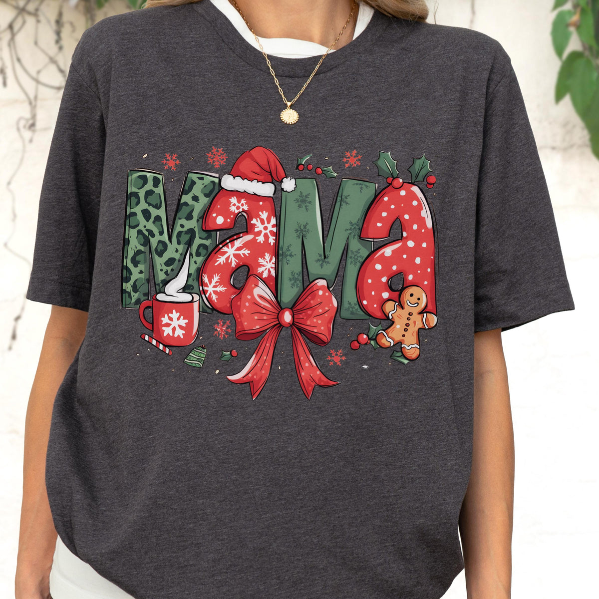 May include: Dark gray t-shirt with a festive "MAMA" graphic. The letters are decorated with Christmas elements: leopard print, snowflakes, a Santa hat, holly, and a gingerbread man. A red bow and a mug of hot cocoa complete the design.