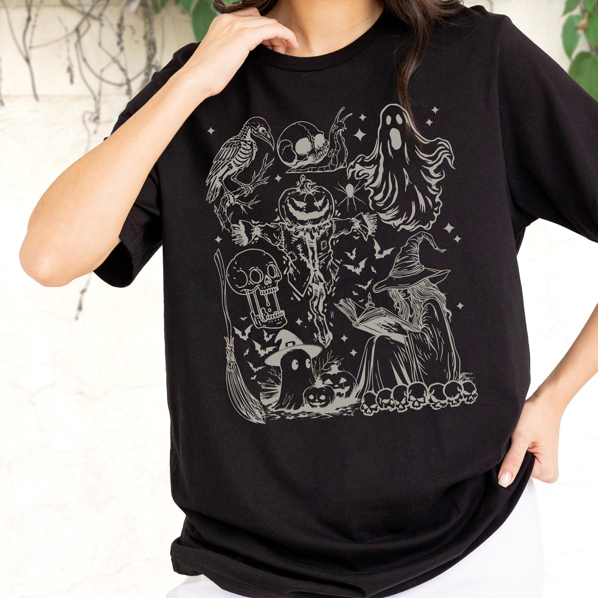 May include: Black t-shirt featuring a detailed, light gray graphic of Halloween-themed illustrations. The design includes a witch, skulls, a ghost, a pumpkin, bats, and a broom. The shirt is made of a soft material.
