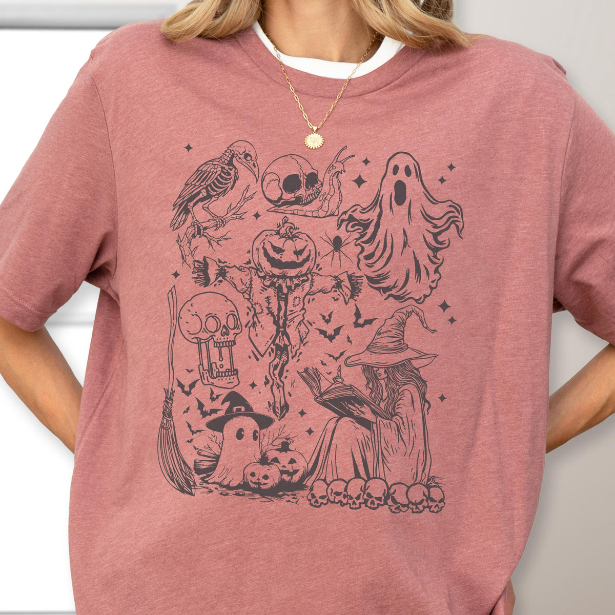 May include: A dusty rose-colored t-shirt featuring a black graphic design of Halloween-themed elements. The design includes a crow skeleton, a skull, a ghost, a scarecrow with a pumpkin head, a witch reading a book, and other spooky elements.