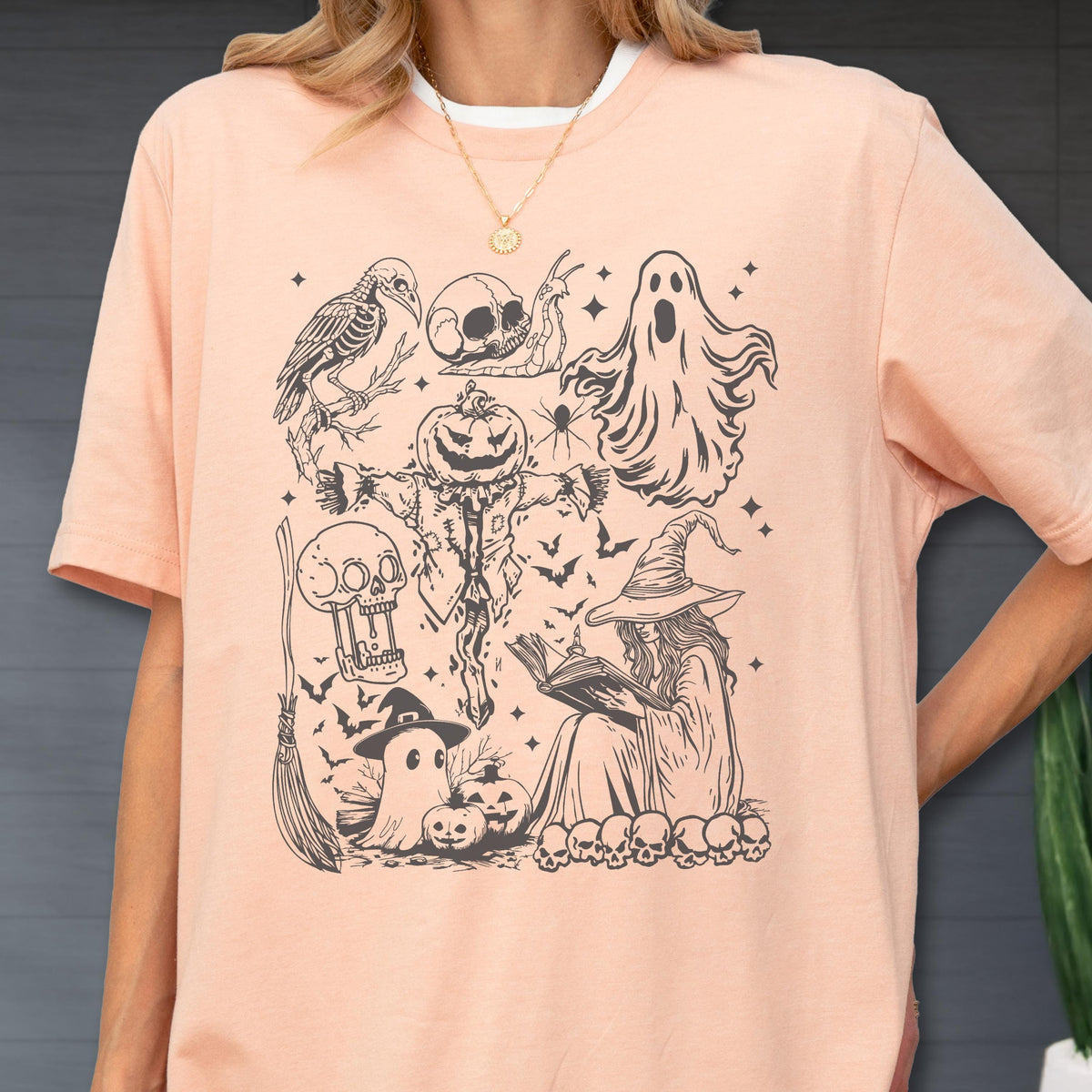 May include: A light peach-colored t-shirt featuring a black and white Halloween-themed graphic. The design includes a witch, ghosts, pumpkins, bats, and other spooky elements. The shirt has a crew neck and short sleeves.