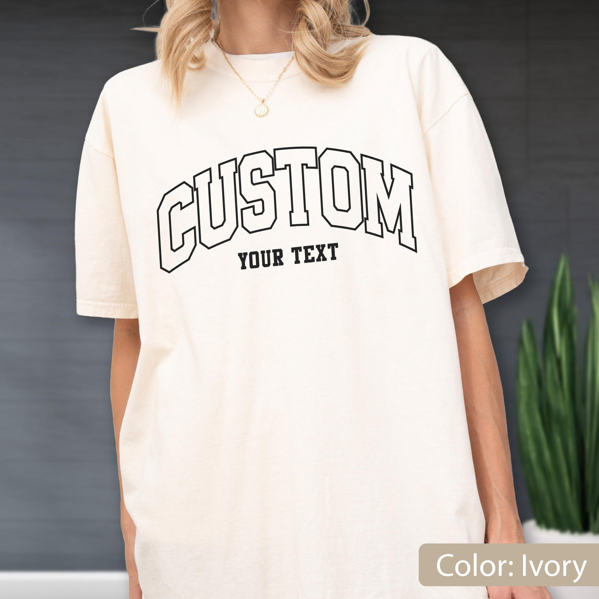May include: Ivory-colored t-shirt with the word "CUSTOM" arched across the chest in a black, outlined font. Below "CUSTOM" is the text "YOUR TEXT" in a smaller, black font. The shirt has short sleeves and a relaxed fit.