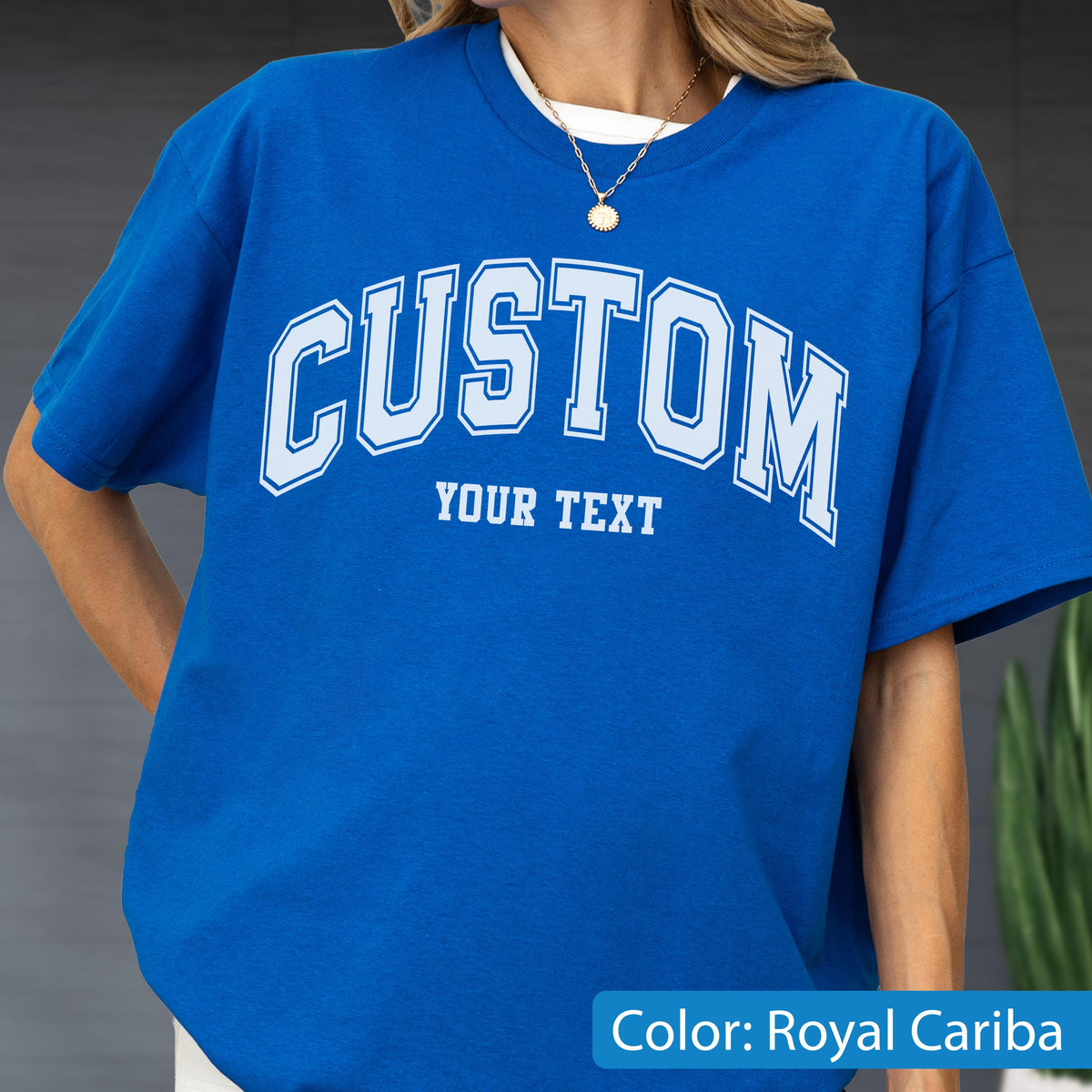 May include: Royal Cariba blue t-shirt with the word "CUSTOM" arched across the chest in white block letters with a light blue outline. Below the word "CUSTOM" is the text "YOUR TEXT". The shirt has short sleeves.