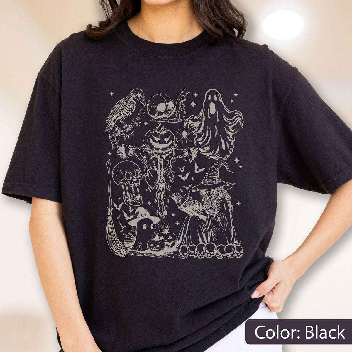 May include: Black t-shirt featuring a detailed, grayscale illustration of Halloween-themed elements. The design includes a crow, skulls, a ghost, a scarecrow with a pumpkin head, a witch reading a book, bats, and other spooky imagery. The text "Color: Black" is at the bottom.